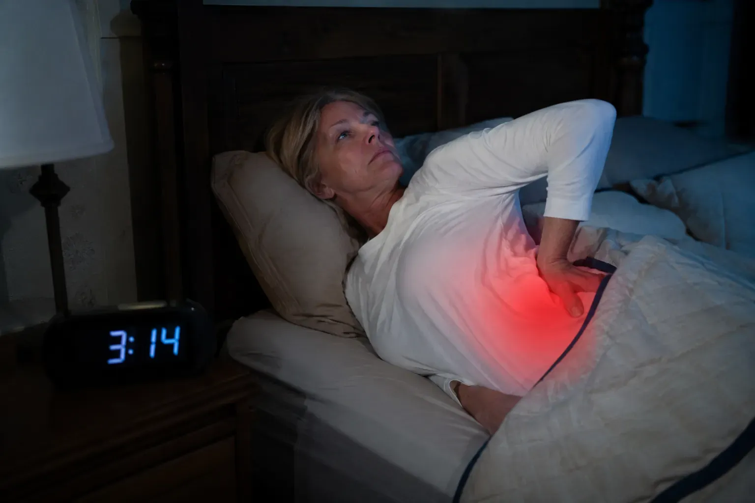 A woman in her early 60s lies awake on her back in bed at night, one hand pressed to her aching lower back, a clock reading 3:14