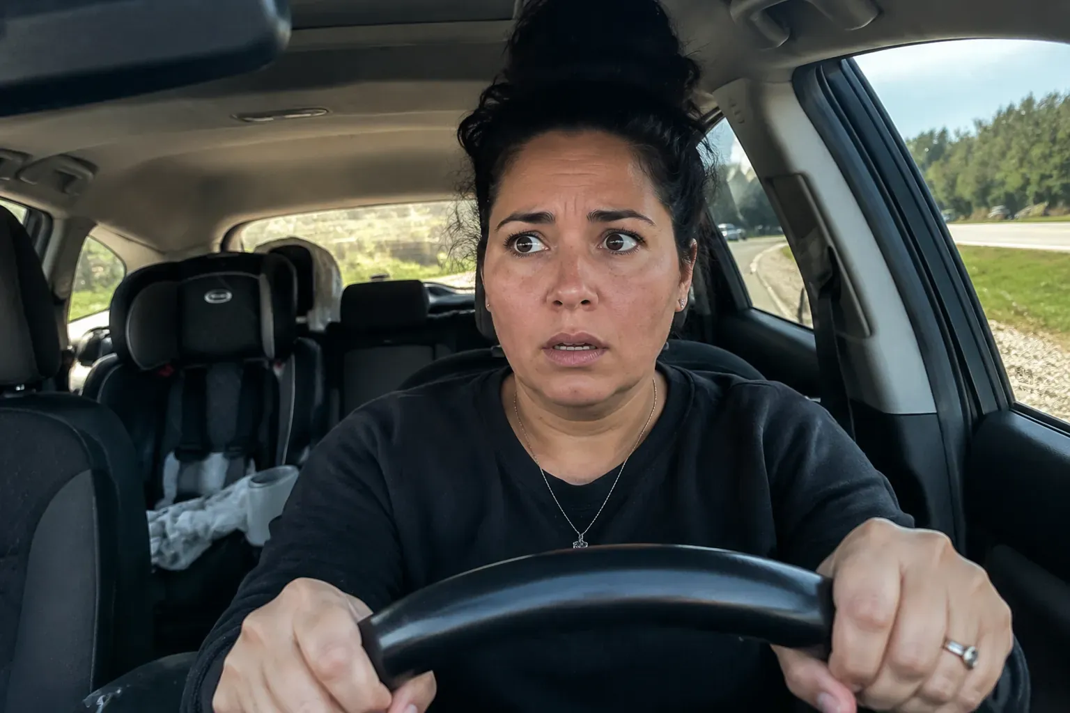 The same woman pulled over on the shoulder of a quiet road, both hands gripping the wheel, shaken, a child's car seat visible in the back